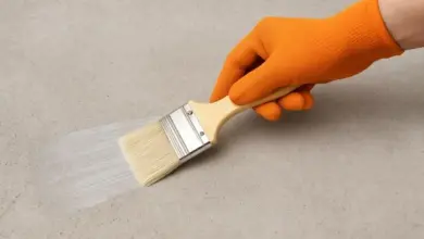 Ruka v rukavici nanáší bílou barvu na štukovou stěnu štětcem.