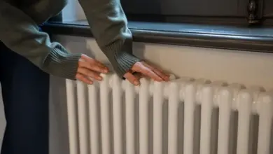 Osoba dotýká bílého radiátoru pod oknem v interiéru.