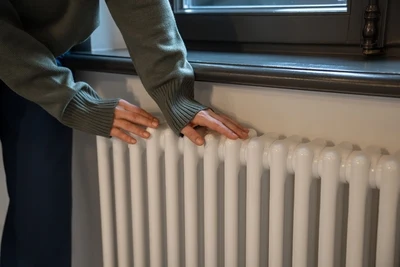 Osoba dotýká bílého radiátoru pod oknem v interiéru.