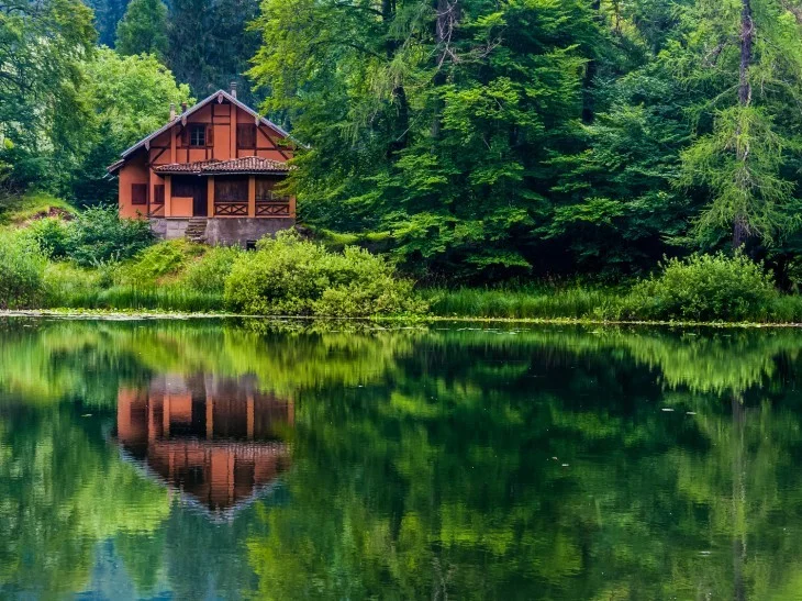Dřevěný domek u jezera v lesu s odrazem ve vodě.