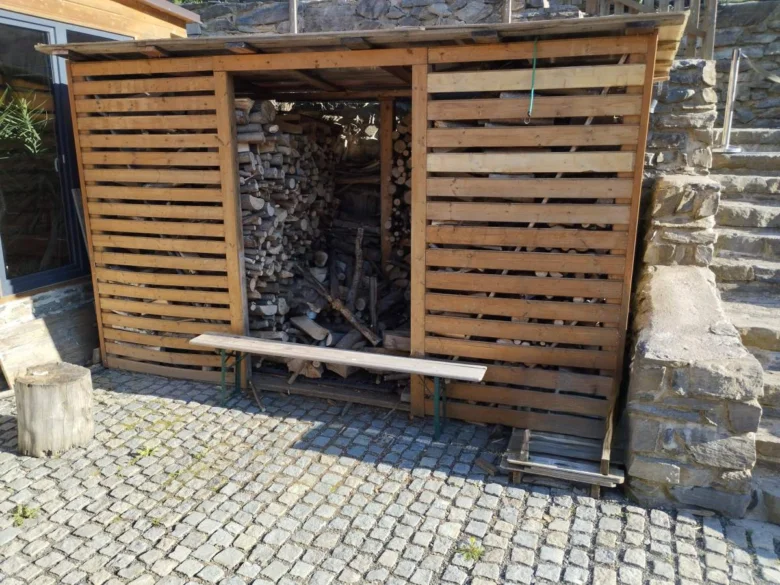 Dřevník s naskládaným dřívím na kamenné dlažbě.
