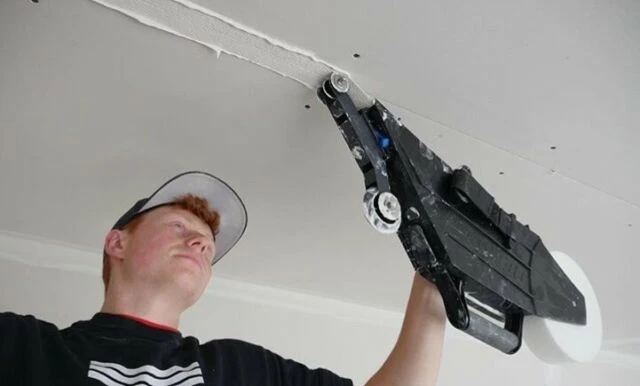 Muž nanáší tmel na švy sádrokartonu stropu pomocí speciálního nástroje.