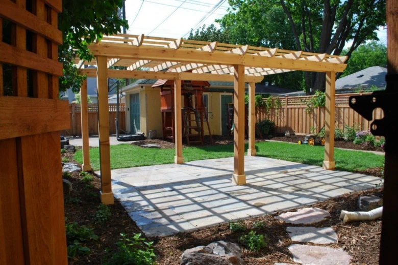 Nová dřevěná pergola s betonovou dlažbou v zahradním pozadí.