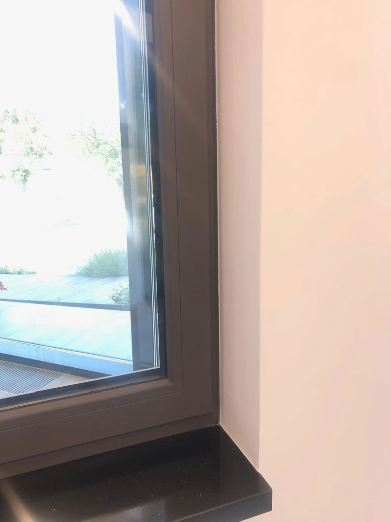 Detail tmavého rámu okna uvnitř místnosti s bílou stěnou.