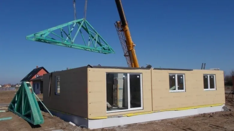 Stavební kran zvedá zelený nosník na střechu prefabrikovaného domu.