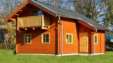Dřevěná chata s balkónem na trávníku před stromy.