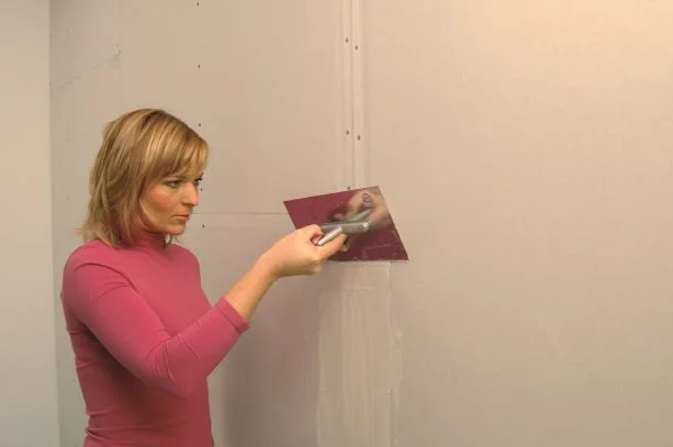 Žena nanáší tmel na spoji sádrokartonu špachtlí.