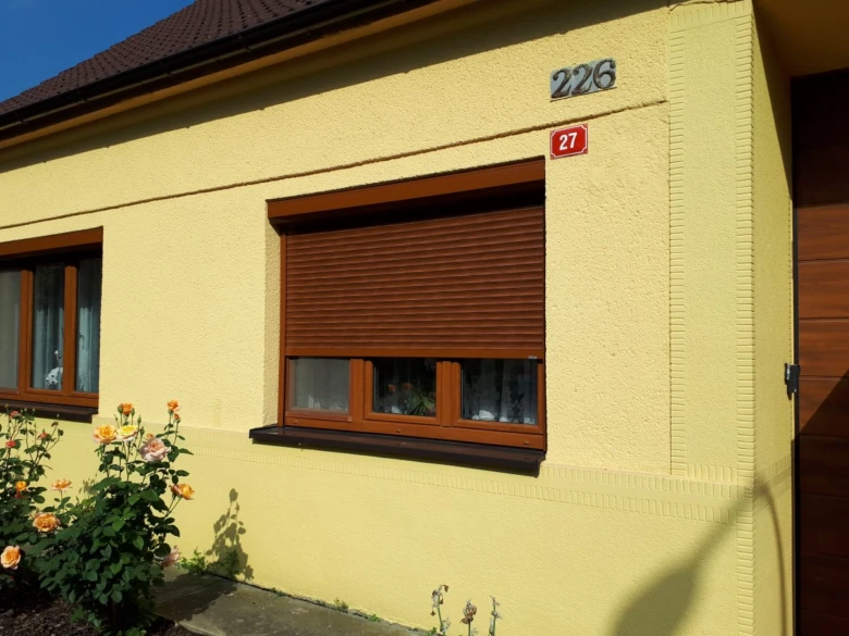 Venkovní žaluzie na okně žlutého domu s kvetoucími růžemi před ním.