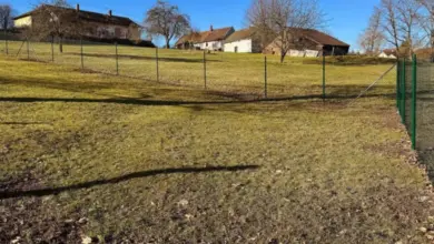Zelený drátěný plot na svahu s domy v pozadí.