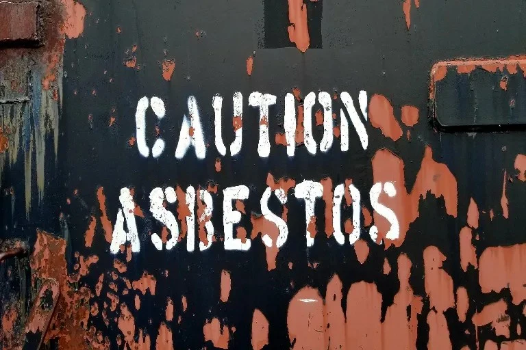 Varovný nápis CAUTION ASBESTOS na zdi s odlupující se barvou.