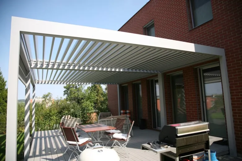 Bílá kovová pergola u červené cihlové domu s venkovním jídelním setem.