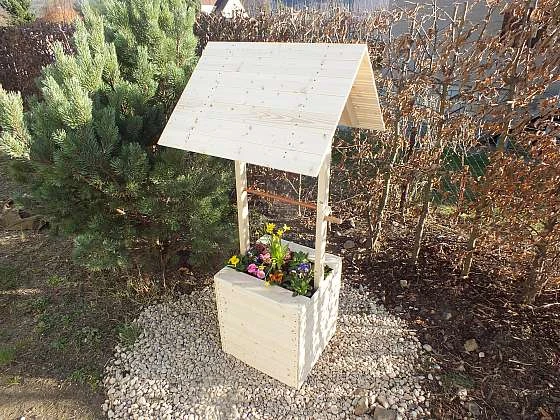 Dřevěný rumpál s kvetoucími rostlinami na zahradním pozadí.