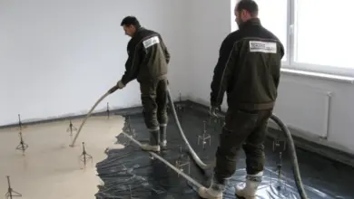 Dva dělníci nalévají podlahovou směs na černou fólii v prázdné místnosti.