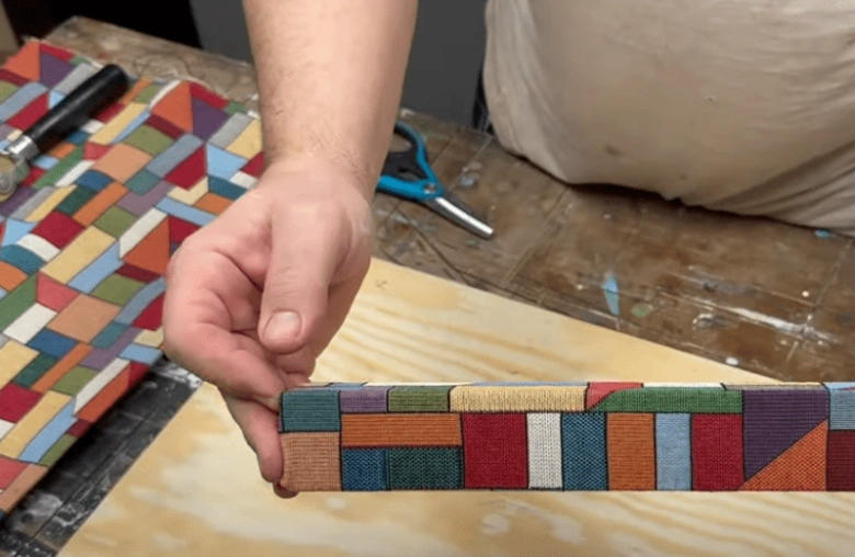 Ruka drží čalouněný rám s barevným geometrickým vzorem na stolu.