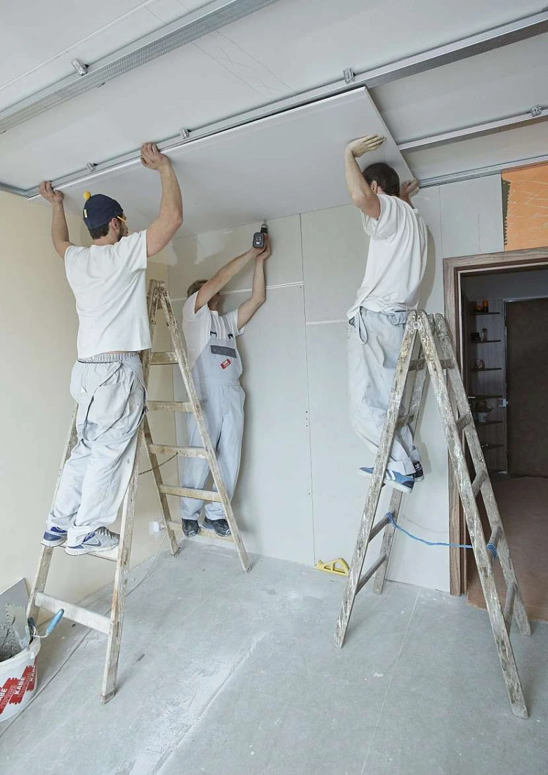 Tři muži montují sádrokartonový strop na lešení.