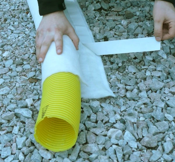 Osoba izoluje žlutou drenážní trubku bílým materiálem na štěrku.