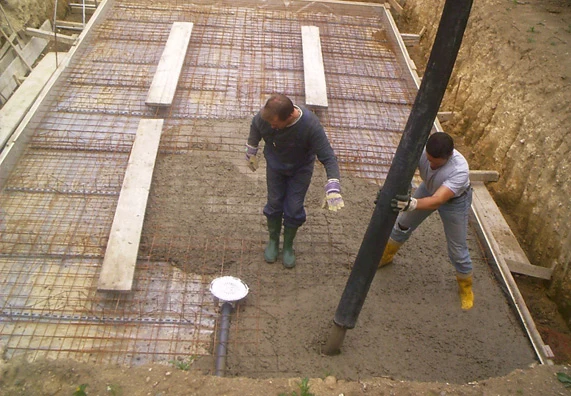 Dva dělníci nalévají beton do základu bazénu s výztuhou.