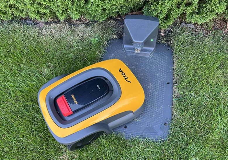 Žlutá robotická sekačka na nabíjecím stanovišti v zahradě.