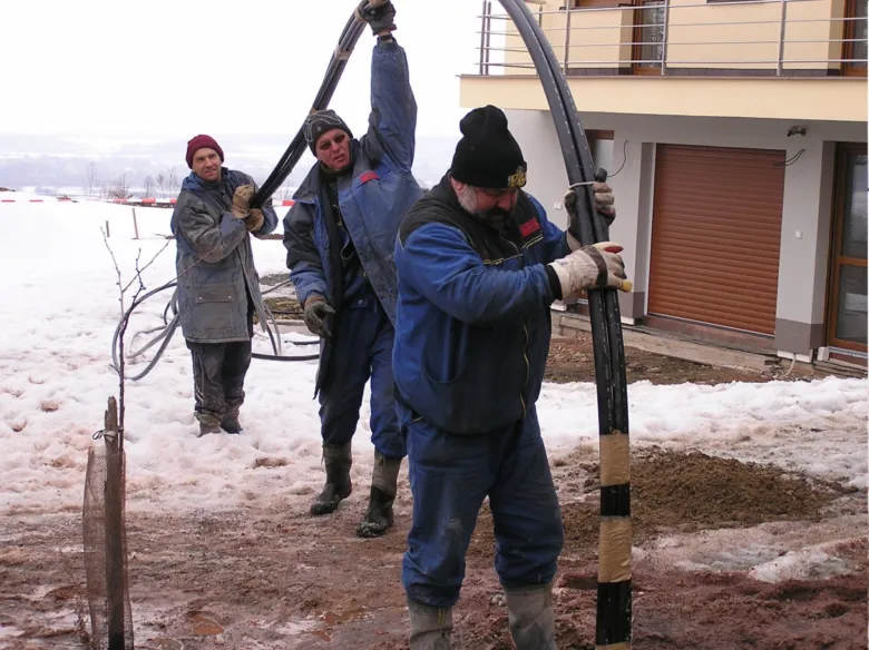 Tři dělníci zvedají velkou černou geotermální trubku na sněhu.