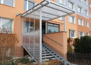Skleněná pergola nad schodištěm k vchodu do budovy.