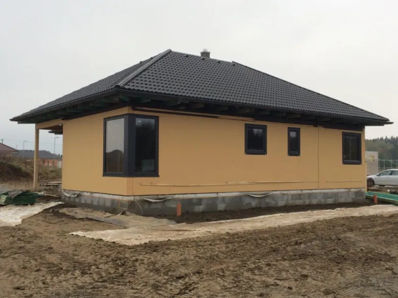 Nově postavený žlutý dům s tmavou střechou na nezpevněném pozemku.