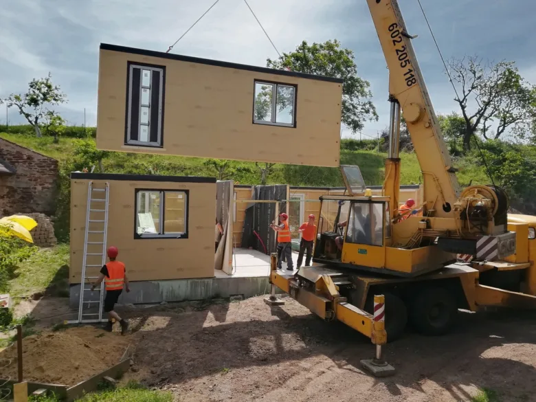 Žlutý kran zvedá modulový dům na základnu s dělníky.